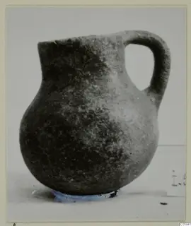 Coarse ware jug with a rounded body, narrow neck and loop handle, photographed on a pale surface.
