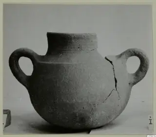Coarse pottery jug with two loop handles, a tall rim and a cracked body, photographed on a plain surface.