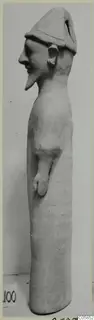 Tall terracotta standing figure with a conical cap, seen from the side against a plain background