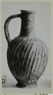 Wheel-made bucchero jug with a single handle and vertically grooved body, photographed on a pale surface