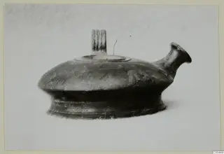 Dark pottery vessel with a spout and a grooved vertical handle on top, photographed on a plain background