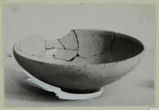 Broken pottery bowl with a rounded body and wide mouth, photographed on a pale surface with a white strip beneath.