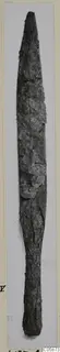 Long corroded iron blade, pointed at the top with a narrow tang at the bottom, shown upright on a pale card mount.