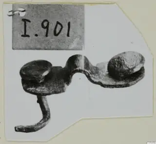 Corroded metal mounting fragment with two domed knobs joined by a curved bar and a bent arm, photographed on a card mount.