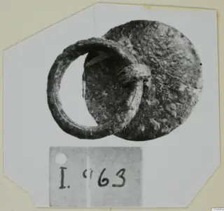 Corroded metal ring attached to a round disc-shaped fragment, with a number label below on a white card