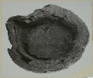 Heavily corroded metal object with a shallow bowl form and broken rim, photographed from above on a pale card.