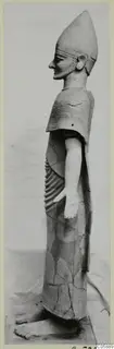Side view of a standing terracotta figure with a tall conical headdress, a short beard and a ribbed cape.