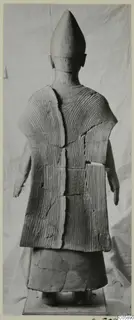 Back view of a terracotta standing figure with a tall conical head and incised parallel lines on its garment