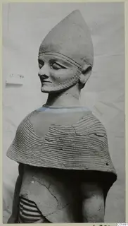 Terracotta statue with a tall conical head covering, braided beard and cape with horizontal incised lines