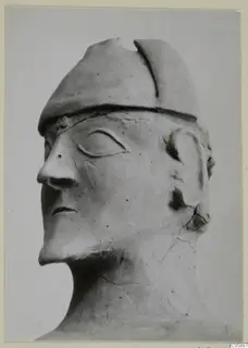 Terracotta head with a cap-like headdress and a long nose, shown in close view against a plain grey backdrop.