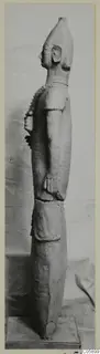 Side view of a tall terracotta statue of a standing figure with a tall head, on a wooden base