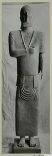 Standing terracotta statue of a figure with a cap and necklace, on a rectangular base, shown against a draped cloth