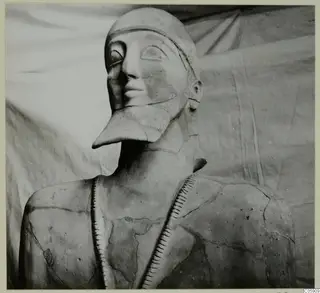 Large terracotta statue of a figure with a long pointed beard and a cap, seen from the chest up against a cloth backdrop.