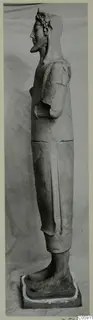 Side view of a tall standing terracotta statue on a square base, with a wreath or headdress and one arm bent at the chest.