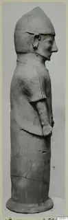 Terracotta statue of a standing figure in side view, with a tall pointed head covering and arms held against the body