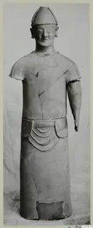 Terracotta standing figure with a tall conical cap and one arm missing, photographed against a draped cloth