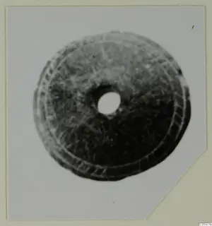 Round disc with a central hole and concentric incised rings, shown in a photograph mounted on a card.