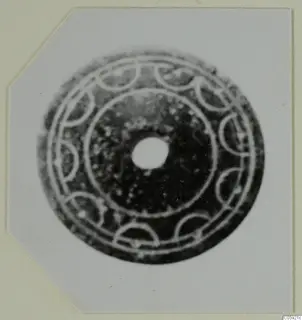 Flat disc-shaped object with a central hole, decorated with concentric rings and a ring of semicircular arcs, shown on a card mount.