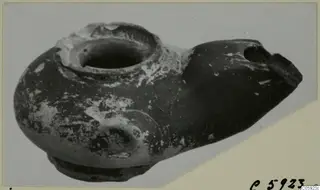 Terracotta oil lamp from Idalion with a broken nozzle, photographed at an angle against a grey backdrop