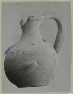 Single-handled pottery jug with a globular body, narrow neck and broken lip, pale surface with dark speckles.