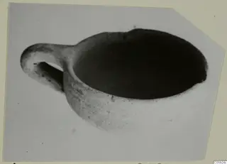Red slip bowl with a horizontal handle, seen from above at an angle against a plain grey backdrop