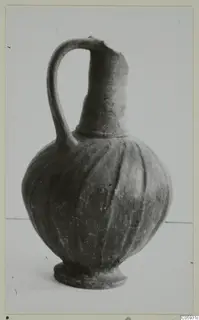 Jug with a single loop handle, tall narrow neck and ribbed globular body, photographed on a plain pale surface.
