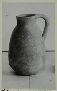 Pottery jug with a single loop handle and a wide mouth, shown on a pale surface against a grey backdrop.