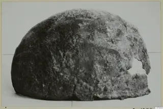 Corroded metal object with a domed shape and a broken opening on one side, photographed against a pale background
