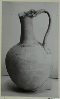 Pottery jug with a tall neck, single loop handle and broken rim, photographed against a grey backdrop.