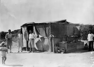 People standing in front of a reed-mat camp shelter at Oura, with a child in a sun hat in the foreground
