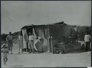 People standing in front of a reed-walled tent at Oura, with a wooden counter beside it.