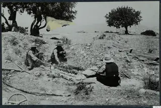 Men sit on a stony slope roasting food on skewers at Vouni, under a tree