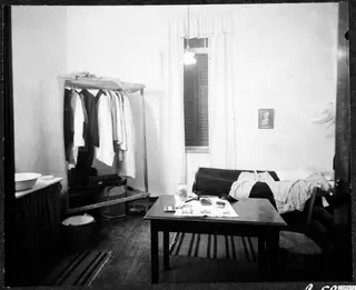 Bedroom in Nicosia with a wardrobe rack of clothes, a table with papers, and a bed under a window.