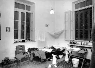 Bedroom in Nicosia with a camp bed, hammock, washstand with basin, and clothing hung on a line.