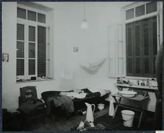 Interior of a room in Nicosia with a camp bed, a folding washstand and tall shuttered windows.