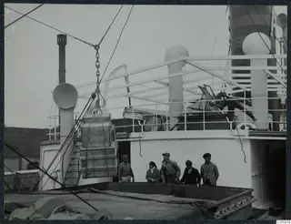 Wooden crates swinging on a chain from a cable on the deck of M/S Gotland at Famagusta, with men and a woman watching.