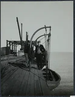 Two men standing on a wooden pier at Karavostasi, with a small boat and the sea beside them.