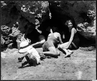Four people at the mouth of a rock cave on a sandy beach, a woman in a dark swimsuit seated at the entrance and a child crouching in front