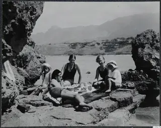 Five people relax and eat on a rocky beach at Kyrenia, with the sea and mountains behind them.