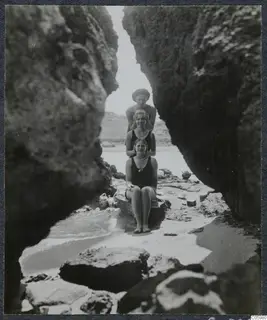 Three women in swimsuits posing on a rock ledge between tall rock walls on a beach, one with hands on the others' heads