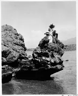 Two people in hats sit on a rocky outcrop above the water on a beach in Kyrenia.