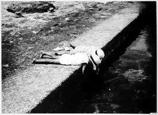 Two children lie on their stomachs on a stone ledge, heads hanging over the water at a beach in Kyrenia