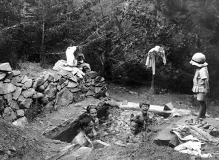 Four people bathing in a stone-lined cistern at Machairas, with a child in a sun hat watching from the edge.