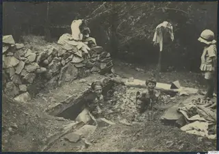 Four people bathing in a stone-lined cistern at Machairas while a child in a hat watches from the edge.
