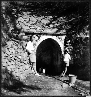 Two people standing beside an arched stone water source, with two jugs and a metal bucket nearby