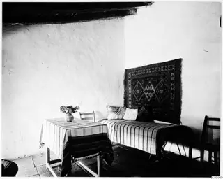 Interior of a room in Machairas with a striped-covered bed, a hanging rug and a table holding a vase of flowers.