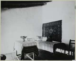 Interior of a whitewashed room in Machairas, with a striped-covered bench, a wall rug and a table holding a vase of flowers.