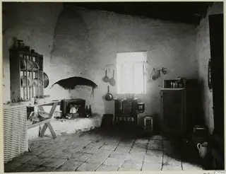 Interior of a house kitchen at Machairas with a whitewashed wall, an arched recess over a stove, and hanging pans.