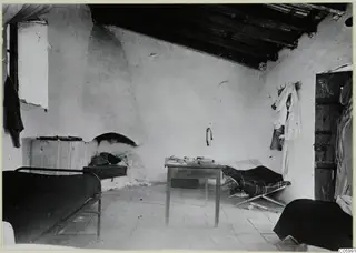 Interior of a whitewashed room at Machairas with two metal beds, a wooden table and an arched recess in the wall.