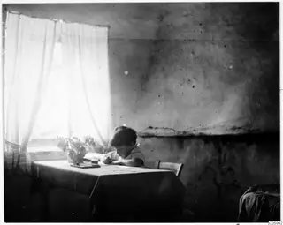 Interior of a house with a small boy writing at a table beside a window, Machairas